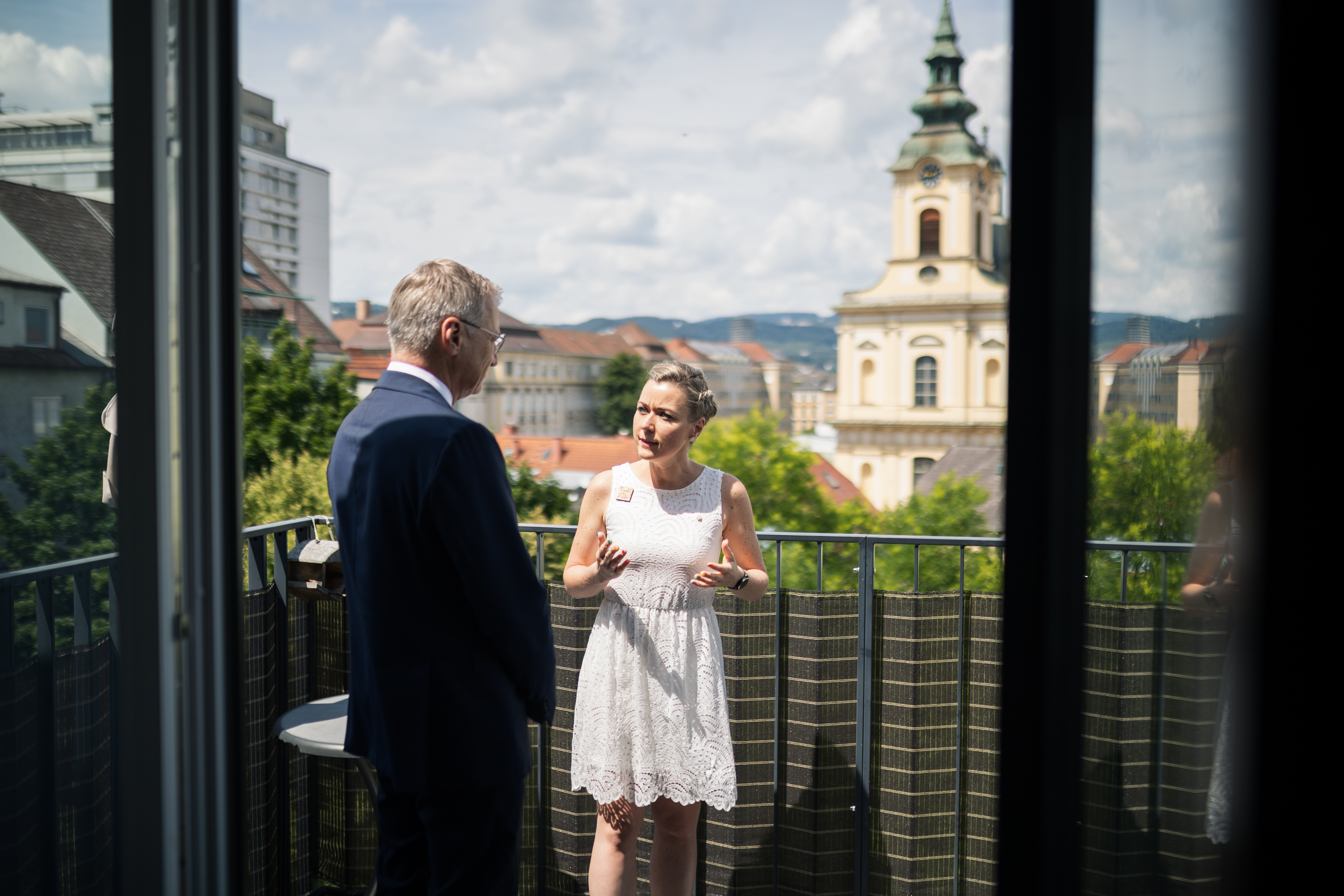 Landeshauptmann im Gespräch / Land OOE Das Bild zeigt den Landeshauptmann von hinten, während er mit einer blonden Frau in einem weißen Kleid spricht. Dabei stehen sie auf einem Balkon.